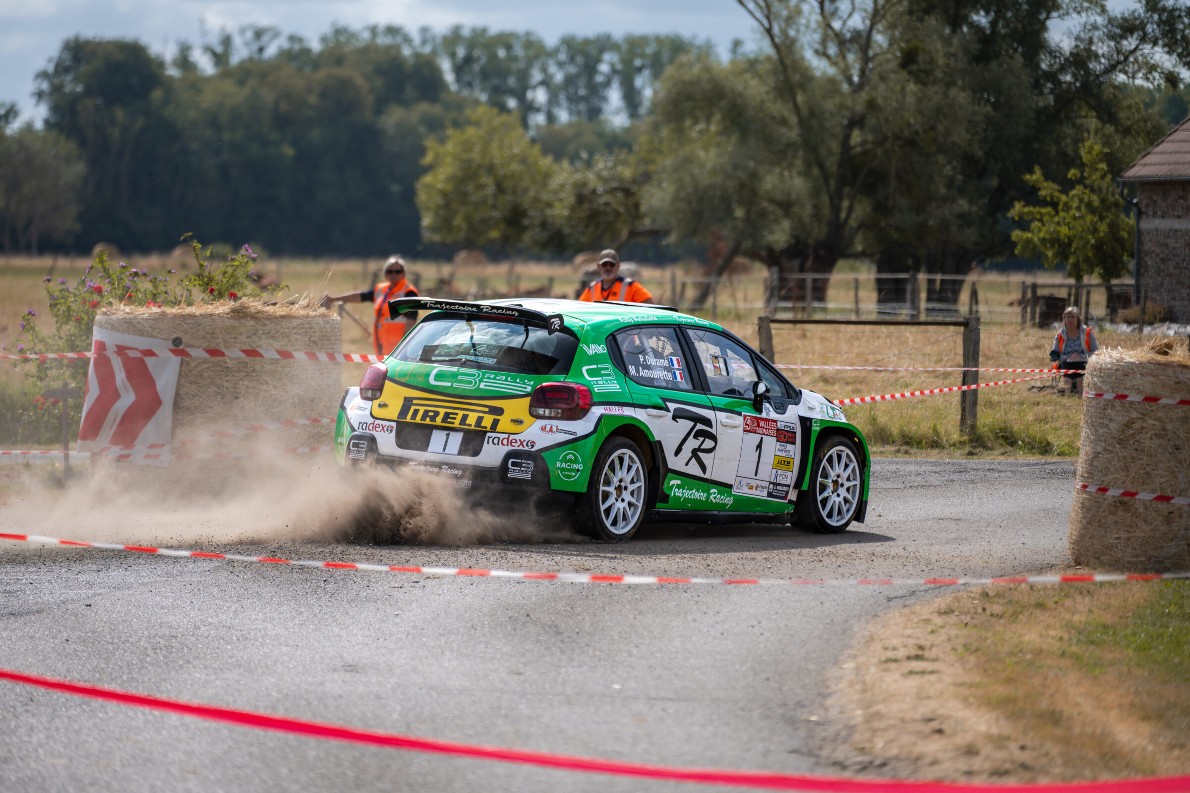Voiture de rallye verte traversant un virage rural derrière une rubalise, avec un nuage de poussière soulevé par les roues arrière.
